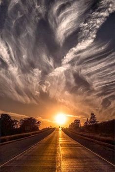 the sun is setting over an empty road with clouds in the sky and trees on either side