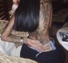 a woman with tattoos on her back sitting in a chair at a table and looking down