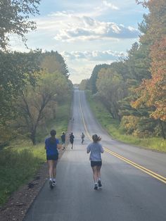 three people running down the road in front of some trees and one person is on roller skates