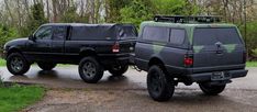 two black trucks are parked on the side of the road near some grass and trees