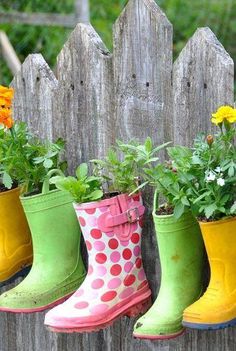 four colorful rain boots are lined up against a wooden fence with flowers growing in them