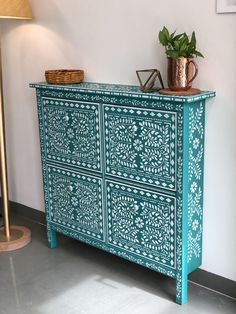 an ornately painted dresser in the corner of a room with a lamp and potted plant on top