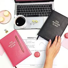 a woman's hands holding a pink notebook next to a laptop and other office supplies