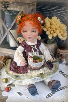 a doll with red hair sitting on a table next to flowers and an old fashioned basket