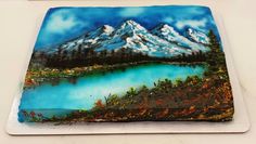 a cake decorated with a painting of a mountain lake and snow capped mountains on it