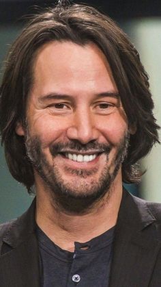 a smiling man with long hair wearing a black shirt and blazer smiles at the camera