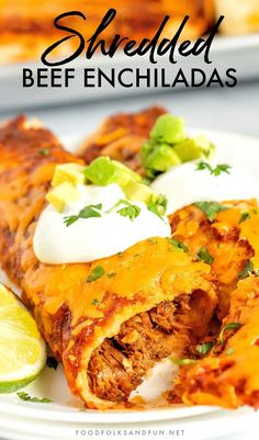 shredded beef enchiladas on a white plate with lime wedges and sour cream