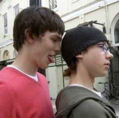 two young men standing next to each other in front of a white building and one is wearing a black beanie