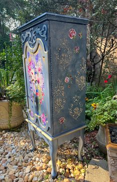 an old blue cabinet with flowers painted on it's doors and drawers is sitting in the gravel