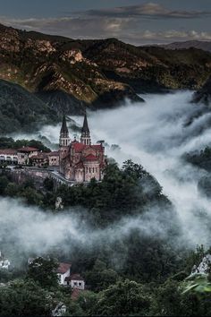 an aerial view of a castle in the middle of trees and mountains, with low lying clouds surrounding it