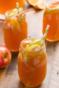 three jars filled with orange juice on top of a wooden table next to sliced apples