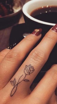 a woman's hand with a small rose tattoo on the middle finger, and a cup of coffee in the background