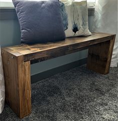 a wooden bench sitting on top of a carpeted floor next to a window with two pillows