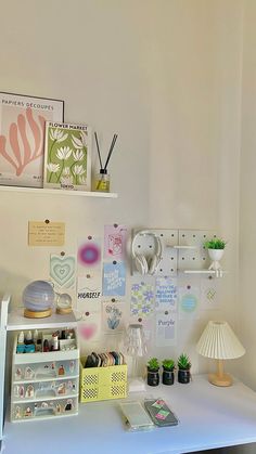 a white desk topped with lots of clutter next to a lamp and pictures on the wall