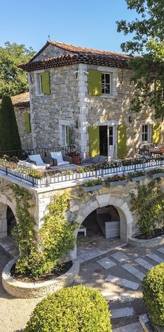 an old stone house with green shutters and trees in the front yard, surrounded by shrubbery