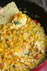 a hand holding a tortilla chip over a skillet filled with corn and cheese