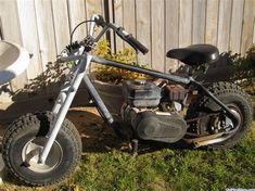 an old dirt bike is parked in the grass next to a wooden fence with a wheelbarrow on it