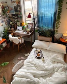 a bedroom with a bed covered in a blanket next to a desk and window filled with potted plants