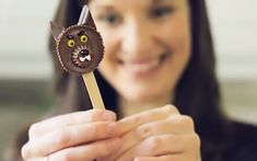 a woman holding up a chocolate cat on a stick with eyes and nose painted on it