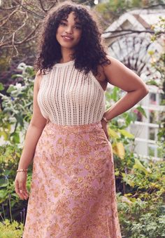 a woman in a pink and gold dress standing next to some bushes with her hands on her hips