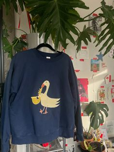 a blue sweatshirt with a yellow duck on it hanging from a rack next to a potted plant