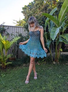 a woman in a blue crochet dress standing on grass next to trees and bushes