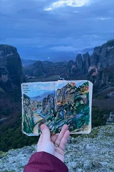 a hand holding an open book on top of a mountain