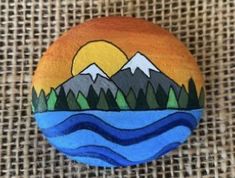 a painted rock sitting on top of a burlock covered canvas background with mountains and trees