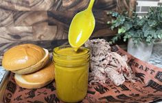 a spoon pouring mustard into a jar next to some bread and buns on a tray