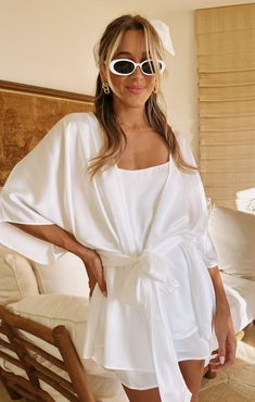 a woman wearing sunglasses and a white dress standing in front of a bed with her hands on her hips