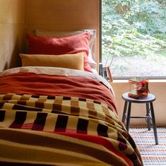 a bed with red and yellow blankets in front of a window next to a small stool