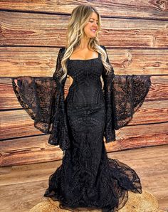 a woman standing in front of a wooden wall wearing a black dress with sheer sleeves