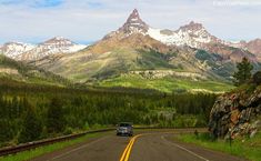 a car is driving down the road in front of mountains with snow capped peaks behind it