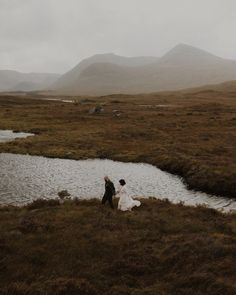 a man and woman walking across a grass covered field next to a river with mountains in the background