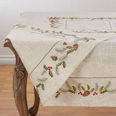 an embroidered tablecloth with holly and pine cones on it, sitting on a wooden bench