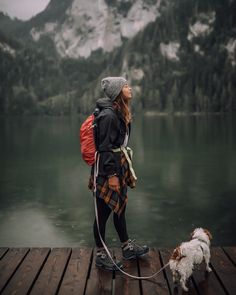 a woman is standing on a dock with her dog and looking up at the mountains