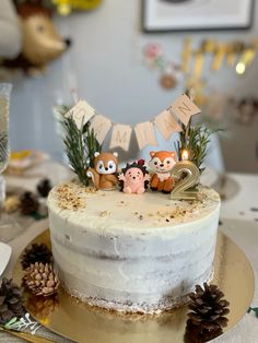 a birthday cake with three little animals on top and pine cones around it, sitting on a table