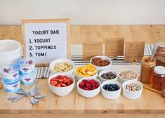 a wooden table topped with bowls of fruit and yogurt next to a sign that says yogurt bar 1 yogurt 2 toppings 3 yum