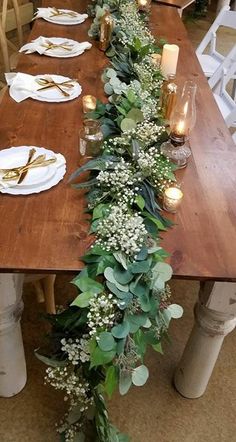 a long wooden table with white plates and candles on it is decorated with greenery