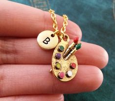 a hand holding a small gold necklace with an artist's palette charm on it