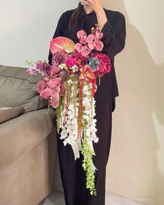 a woman standing next to a couch holding a cell phone up to her face with flowers in front of her