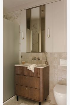 a white toilet sitting next to a bathroom sink under a mirror on top of a wooden cabinet