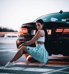 a woman sitting on the ground in front of a car wearing a skirt and sneakers