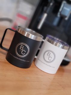 two coffee mugs sitting next to each other on a wooden table with an espresso machine in the background