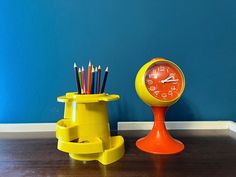 an orange clock next to a yellow cup with pencils in it on a wooden floor