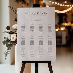 a wedding seating chart on a easel with flowers and lights in the back ground