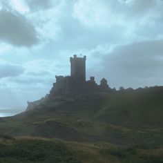 a castle sitting on top of a lush green hillside next to the ocean in front of a cloudy sky