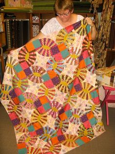 a woman holding up a colorful quilt in front of a bookshelf