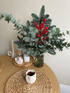 a wooden table topped with a vase filled with red berries and greenery next to a cup of coffee