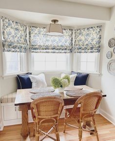 a dining room table with four chairs and a bench in front of the window that has blue and white roman shades on it